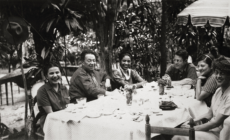 Svartvitt foto på konstnärerna Frida Kahlo och Diego Rivera med vänner sittandes runt ett stort bord. 