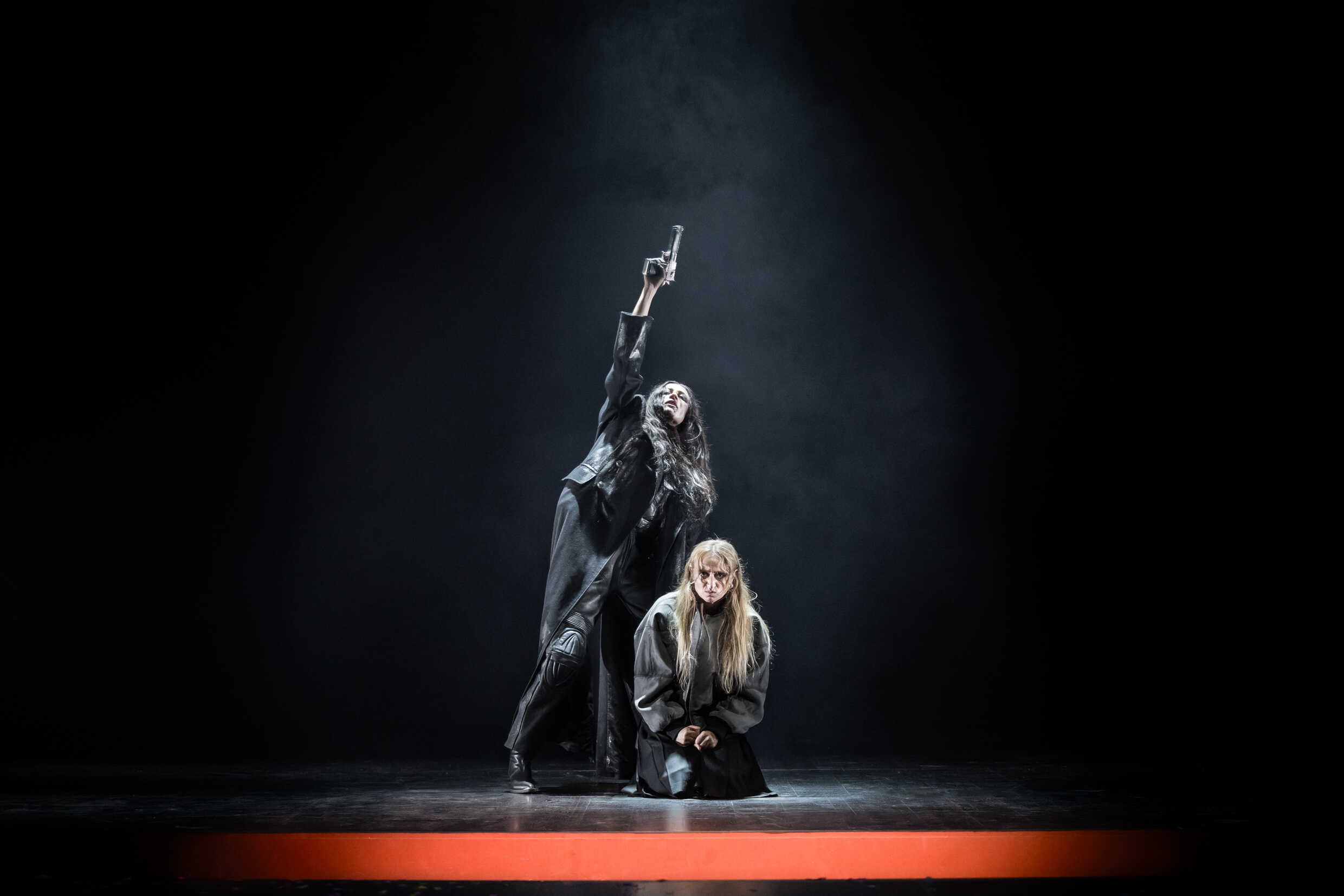 Två kvinnor klädda i svart på en scen, en håller i en pistol. 