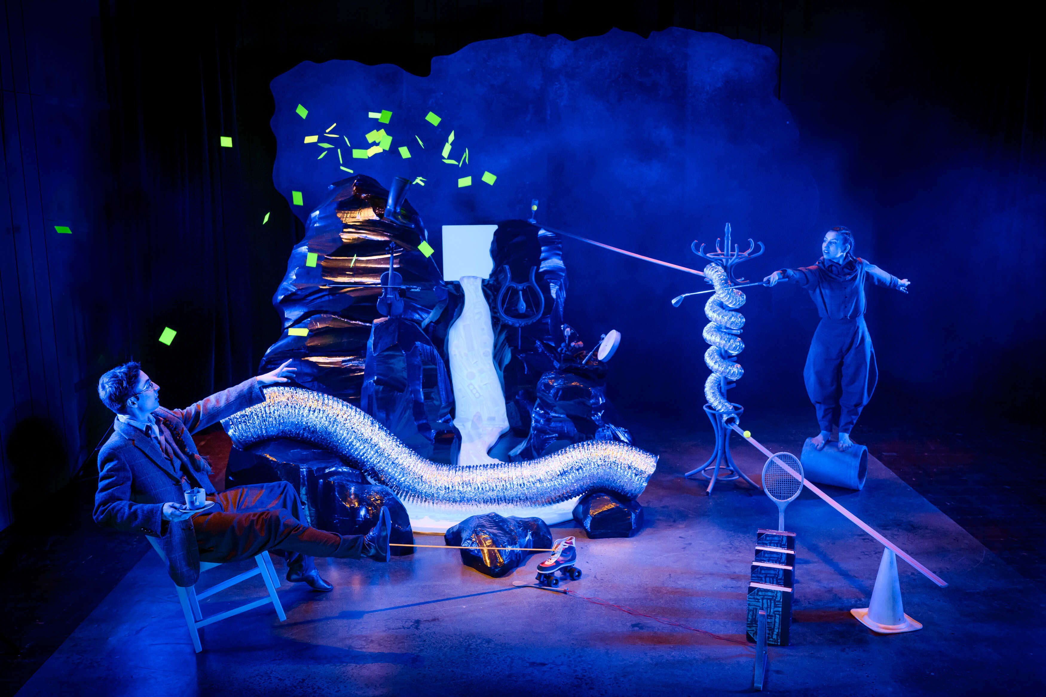 Föreställningsbild från föreställningen Goldbergvariationerna. Ett vattenfall rinner genom dimma och blått ljus. Två cirkusartister är på vardera sida och pekar mot varandra med föremål i händerna