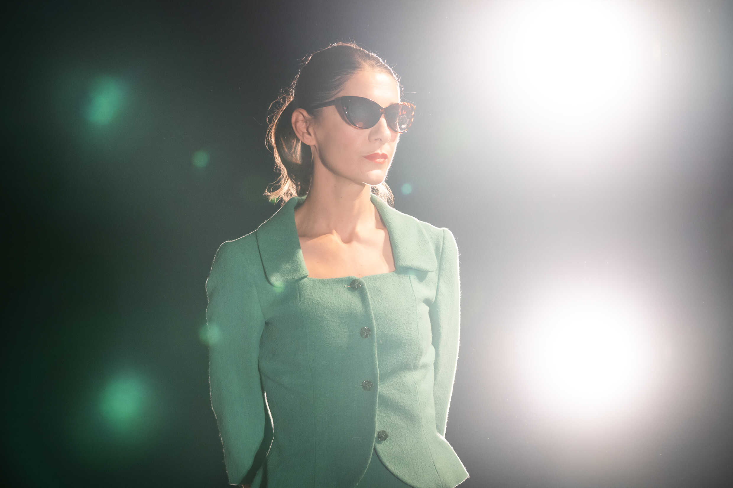 En kvinna i grön dräkt och solglasögon står på en upplyst scen. 