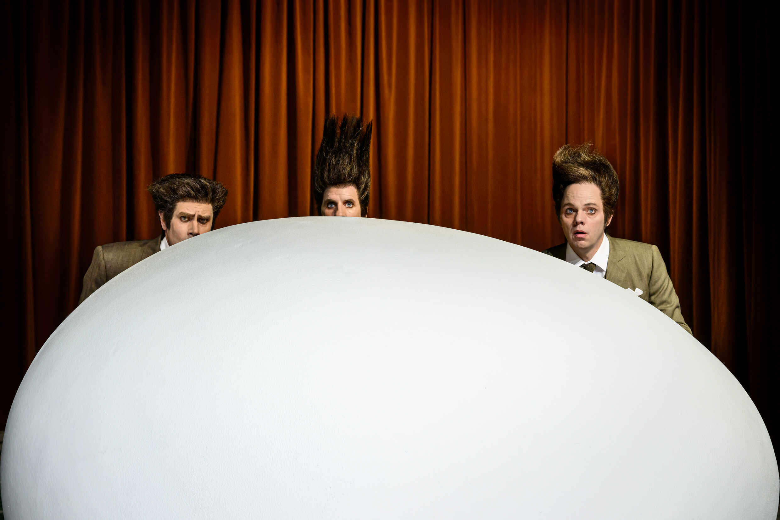 Tre skådespelare bakom en stor vit oval form. 