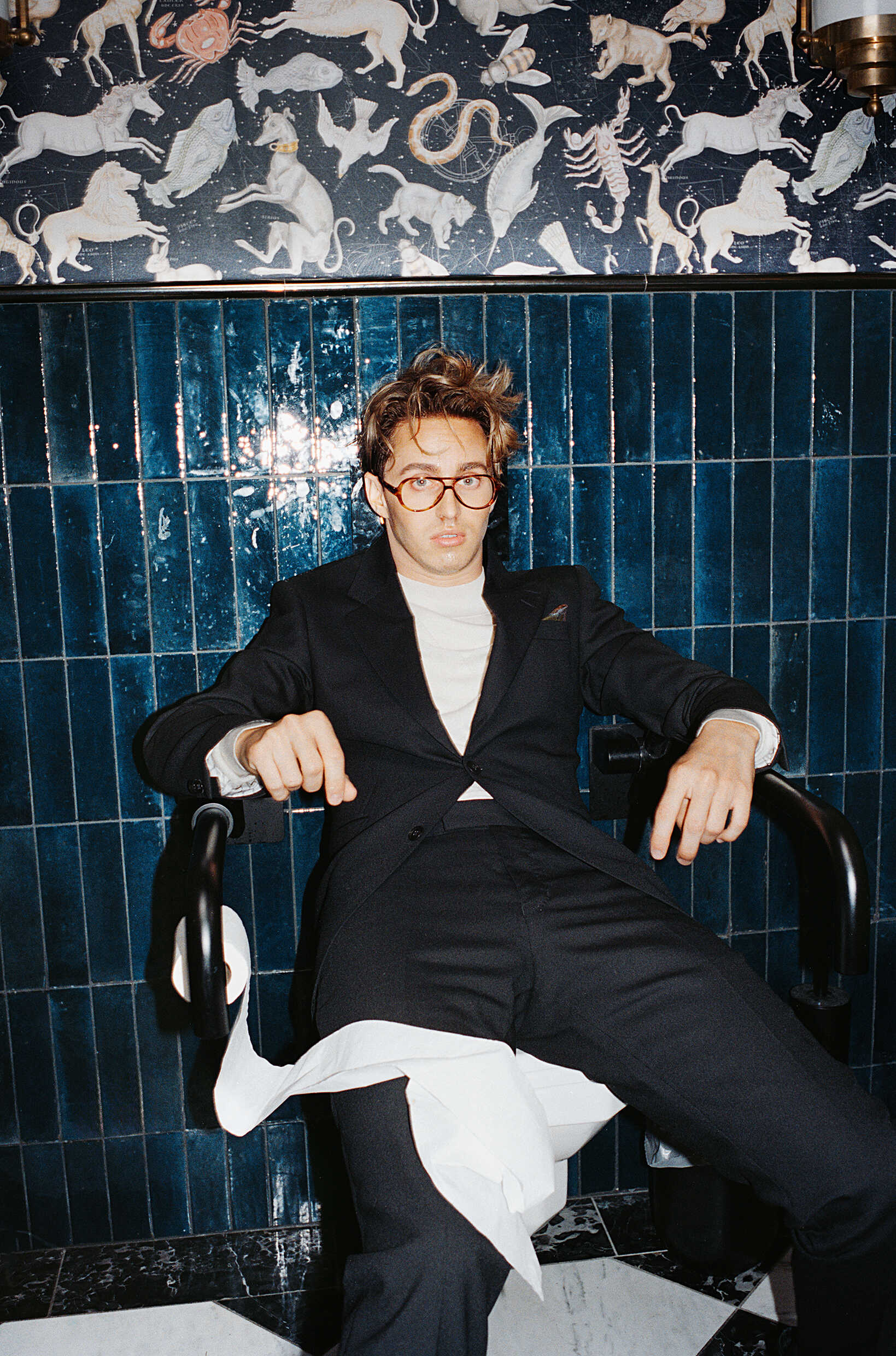 En ung man klädd i kostym sitter på en toalettstol. 