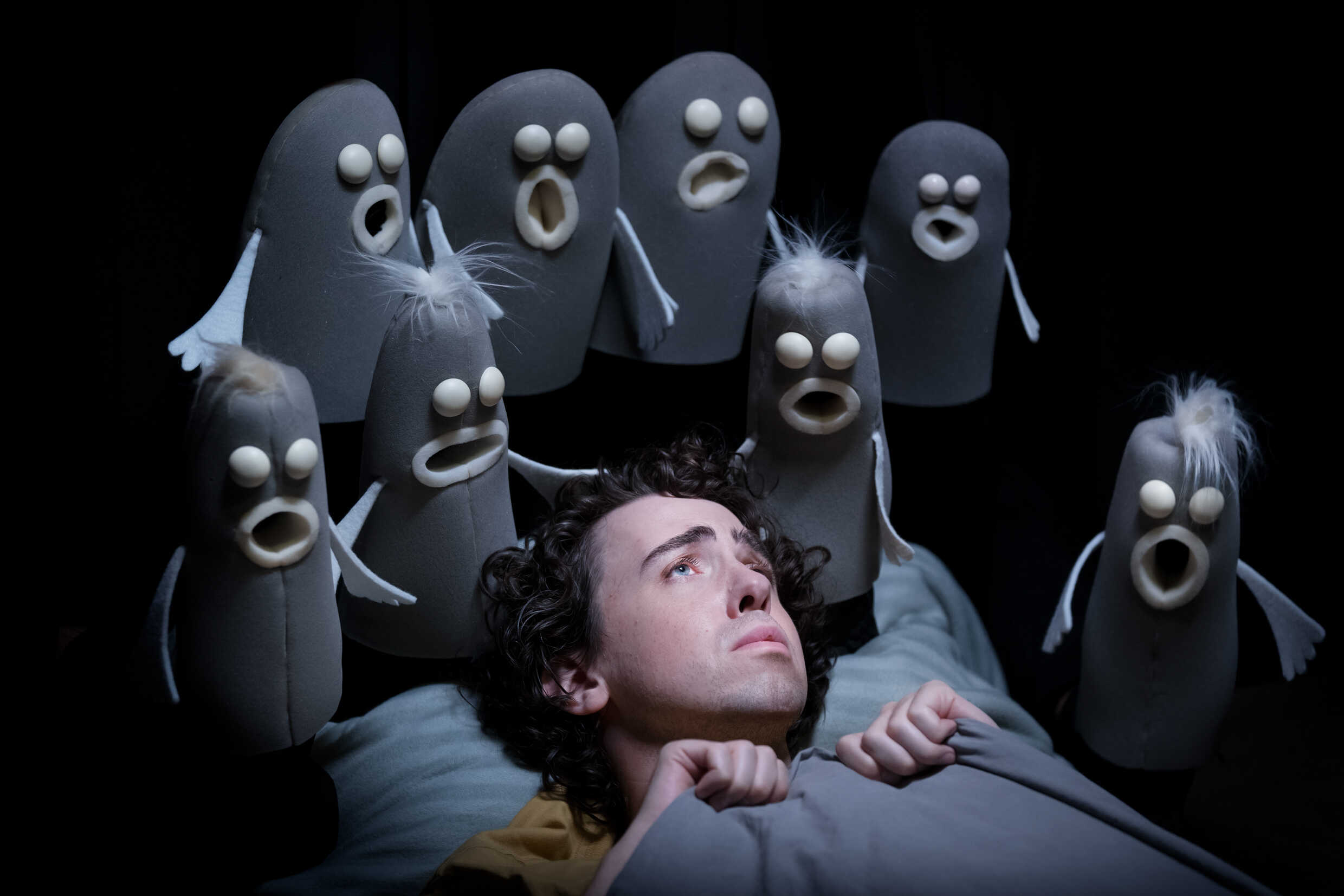 En människa ligger i en säng, omgiven av svarta och grå spökliknande dockor. 