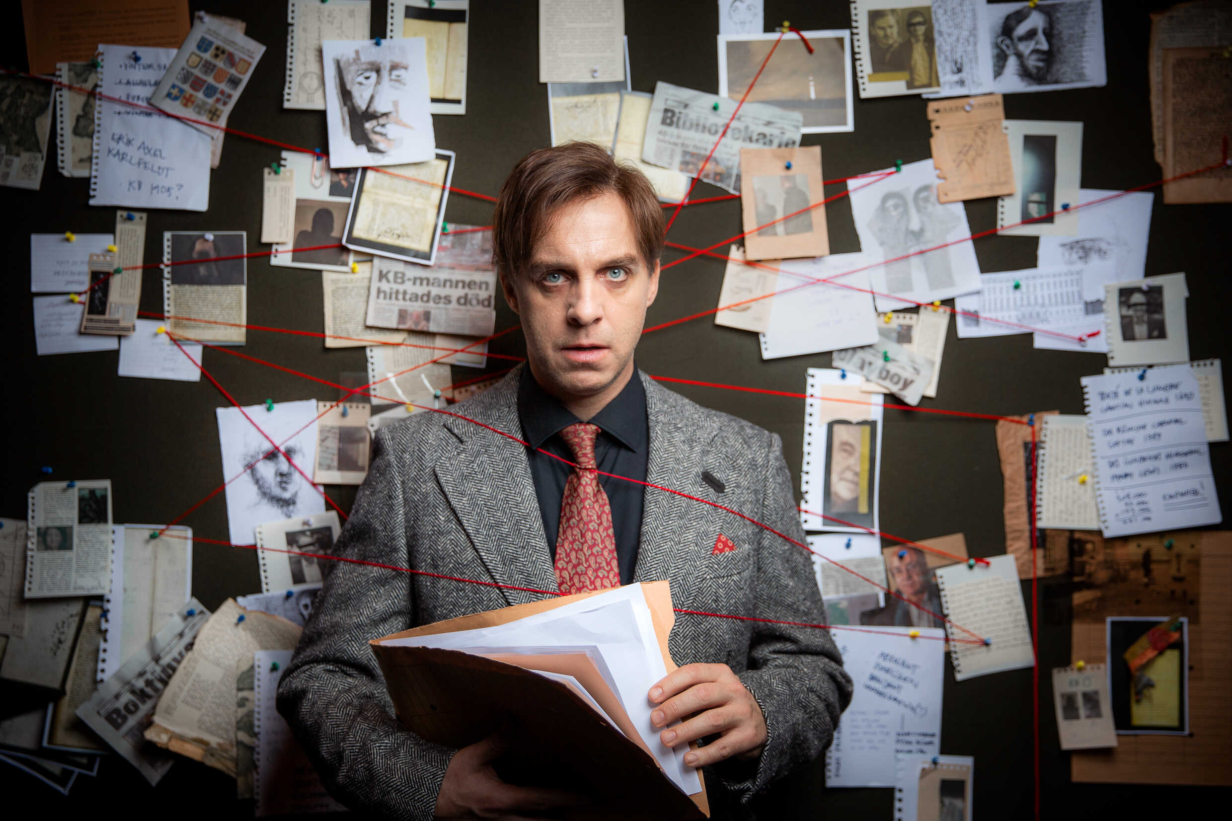 Fredrik Meyer i kostym med en folder i handen. Han står framför en tavla med tidningsurklipp och bilder